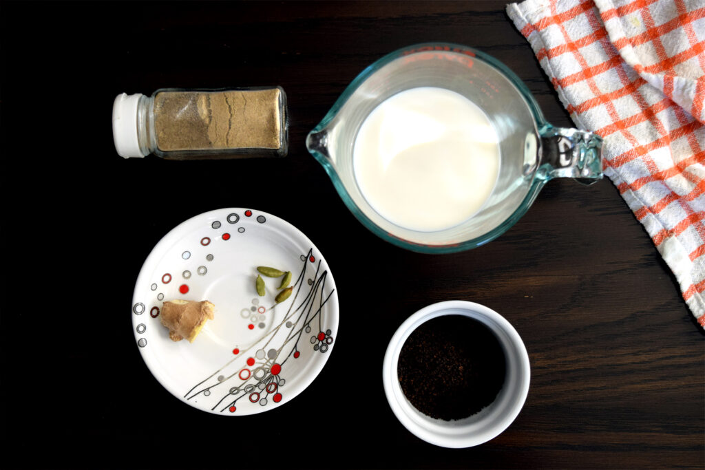 An overhead shot of the ingredients to make chai, including chai masala, ginger, cardamom, tea leaves, and milk. 