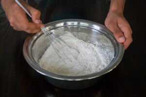 A person whisking flour in a large metal bowl